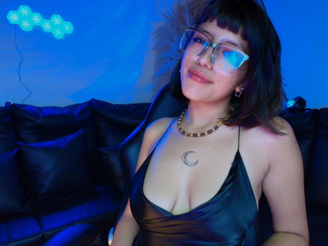 CuteH with glasses, black bob, and a crescent tattoo on her chest, wearing a black halter top, sitting on a black couch, blue light, smiling.