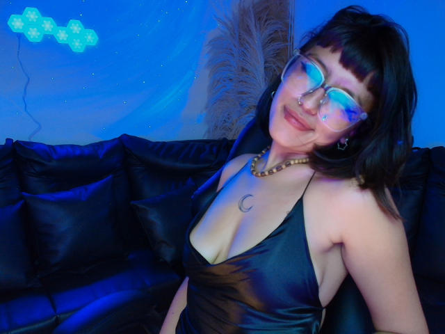 CuteH with blue-tinted glasses, black bob, nose ring, and bead necklace, wearing a black satin dress with deep cleavage, sitting on a black leather couch.