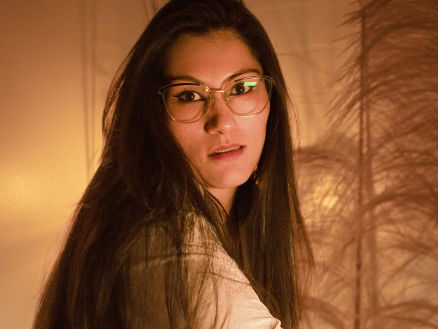RapZelly, con cabello largo y oscuro, gafas y una camisa blanca, mirando por encima del hombro con una ligera sonrisa, iluminación naranja suave y una pluma esponjosa en el fondo..