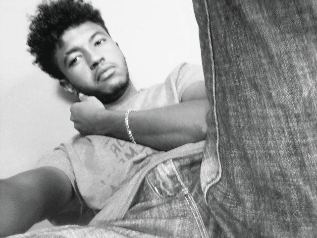Black-and-white selfie of Dacth69 with curly hair, light skin, and a beard, wearing a tee and jeans, arms crossed, bracelet on wrist, legs in the foreground.