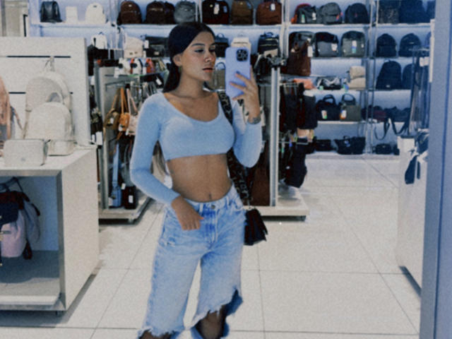 ThaliaAres with taut, toned abs, wearing a light blue crop top, high-waisted, frayed denim pants, and a black headband, taking a mirror selfie in a store.