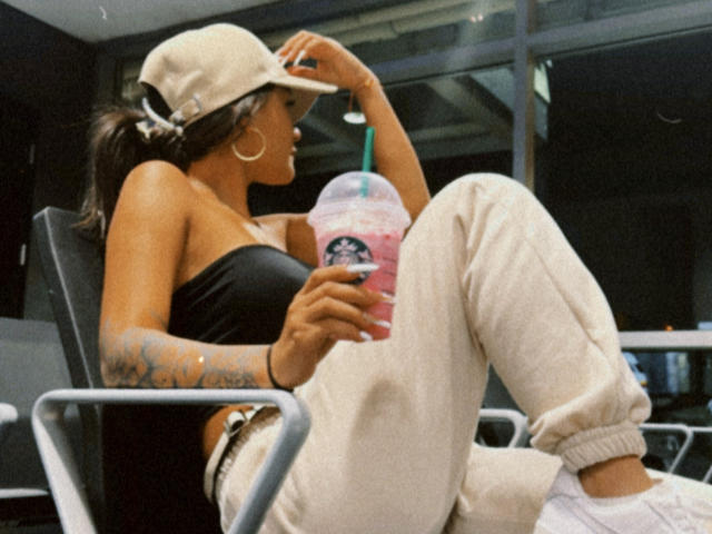 ThaliaAres with tan skin, tattoos on her right arm, black strap top, beige cap, beige pants, holding pink Starbucks cup, sitting in chair, legs crossed, relaxed.