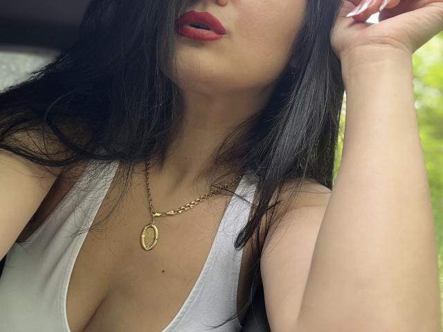 JennaEasy with black hair, red lips, white top, showing cleavage, gold necklace, hand in hair, looking slightly up. Boobs visible, smooth skin, subtle makeup.
