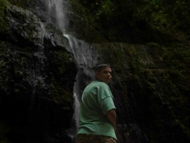 AlexaSandoval with a green shirt and camouflage pants, standing in front of a dark, mossy waterfall, looking back over her shoulder.