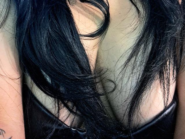 El cabello negro de Viviana Thompson cae sobre su escote, vistiendo una ajustada blusa de cuero negro, mostrando sus grandes, desnudas y pálidas pechos, con piel visible y un pequeño tatuaje en su brazo..