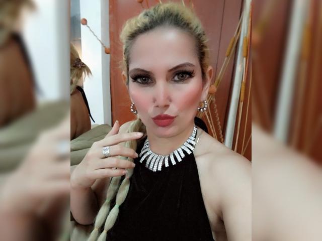 GiuliaAddams a les cheveux blonds, un maquillage audacieux et du rouge à lèvres rouge. Elle porte un haut noir à encolure dégagée, un collier de diamants et des boucles d'oreilles, et une bague à sa main droite..