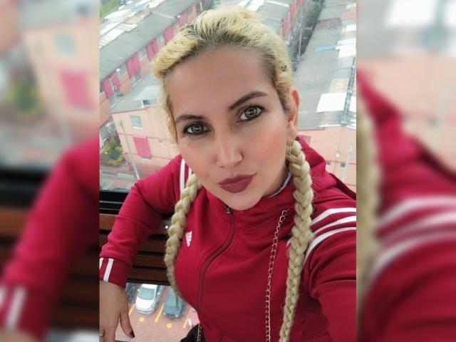 GiuliaAddams con trenzas rubias, chaqueta roja de Adidas, labios fruncidos, mirando hacia la cámara, reflejo en la ventana, vista urbana desde la azotea. Sus pechos son apenas visibles debajo de la chaqueta..