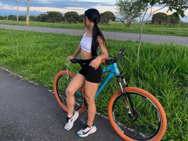 LaurentRay, met lang zwart haar, een witte sportbeha, zwarte shorts en witte sneakers, staat naast een blauwe mountainbike met oranje banden aan de kant van een grasveldige weg..