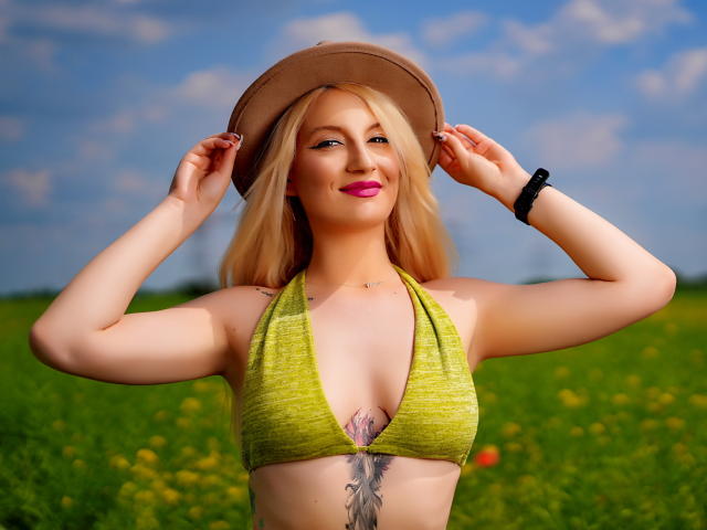 EllaCharm with blonde hair, pink lipstick, green halter top, brown hat, tattoo under boobs, black wristband, smiling, hands on hat, blue sky, green field, bright sunlight.