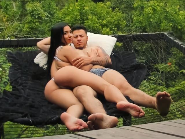 SexyCoupple with black hair, big ass, and tits in white bra, cuddles with tattooed SexyCoupple in blue boxer shorts on black hammock, wood deck, green background.