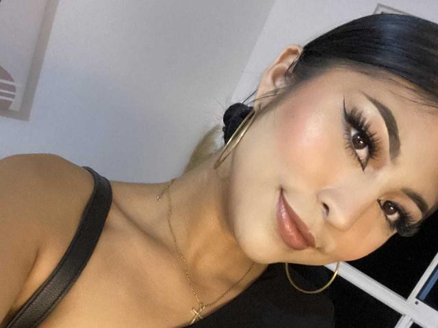 CarlaRusso has dark hair, brown eyes, and a cute smile. She's wearing black, showing her small tits and gold hoop earrings, with a gold necklace. She's looking at the camera.