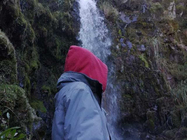SopphiesWett, portant une capuche rouge et une veste bleue, se tient devant une cascade, l'eau ruisselant sur un terrain rocheux et recouvert de mousse. Le dos de SopphiesWett est tourné vers la caméra..