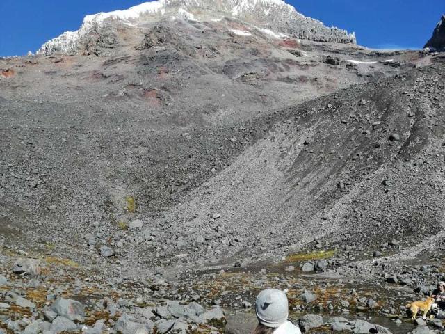 Ein Kopf von SopphiesWett in einem weißen Hut, ein grauer, felsiger Berg mit roten und gelben Flecken, Schnee auf der Spitze, ein klarer, blauer Himmel und verstreute Felsen im Vordergrund..
