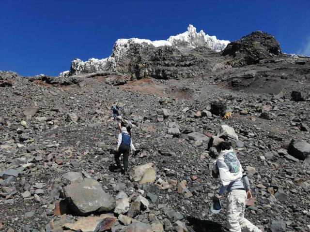 Zwei Bergsteigerinnen, gekleidet in weiße und graue Berbekleidung, besteigen einen felsigen, schneebedeckten Berg unter einem klaren, blauen Himmel. Eine befindet sich vorne, die andere rechts davon..