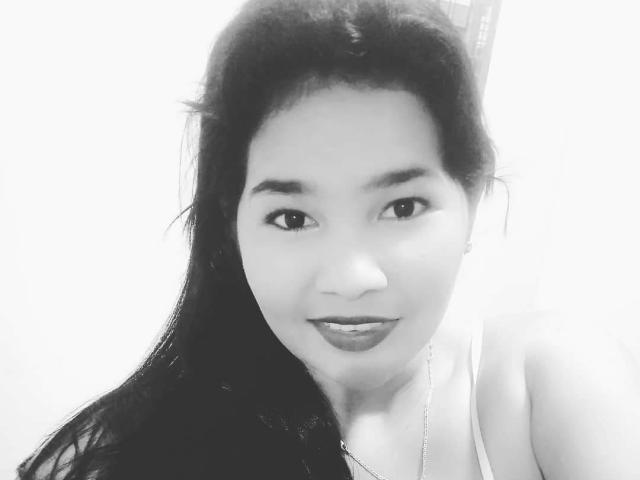 Black and white selfie of PrittyAlicia with long dark hair, medium skin, subtle smile, and dark lipstick. She's wearing a white tank top, showing her small tits.