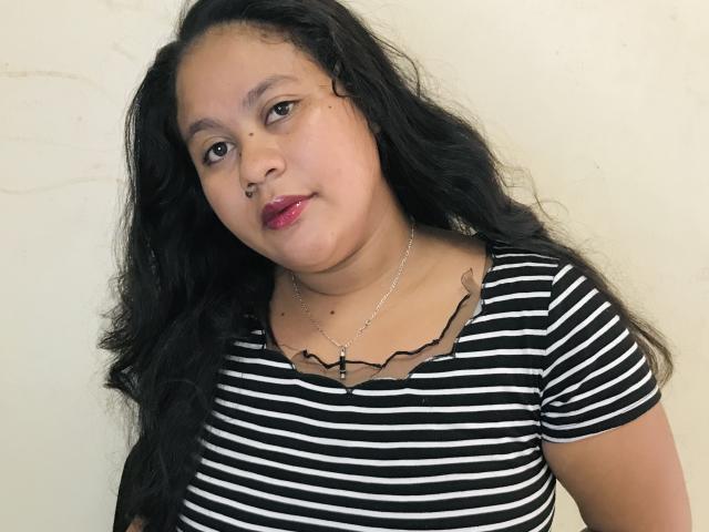 KaityMagik with long black wavy hair, brown eyes, red lipstick, wearing a black and white striped shirt, and a silver necklace, smiling subtly. Her tits are small, and she's cute.