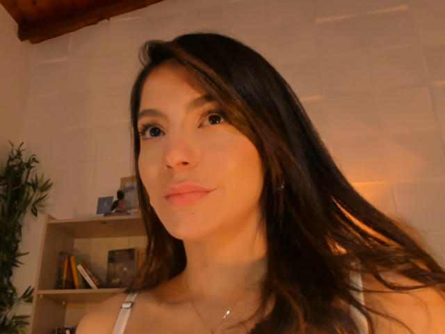 ValenRosee with long brown hair, light skin, and subtle makeup, pouting slightly, wearing a white top, soft light on her face, small gold necklace, gentle smile, warm indoor setting.