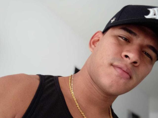 JeicodLatino, avec un léger sourire, portant une casquette noire, un débardeur noir et une chaîne en or, a une corpulence moyenne et une peau brun clair, et regarde de côté..