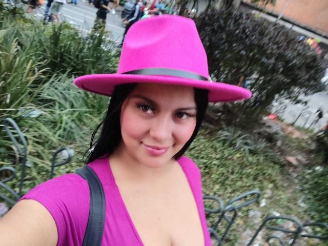 Camihotboobs with pink hat, black hair, pink top, showing cleavage, black strap, smiling, green plants, street background, brown eyes, medium skin, slight makeup, casual selfie.