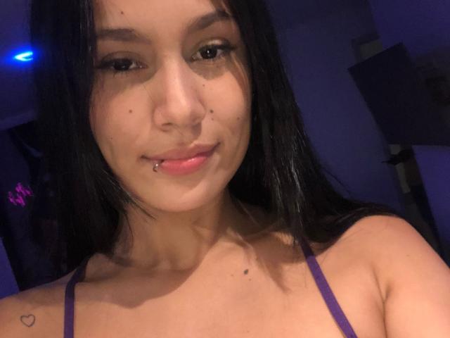 KataleyaMansson has long black hair, a nose piercing, a lip piercing, and a heart tattoo on her right shoulder. She's wearing a purple top, looking cute.