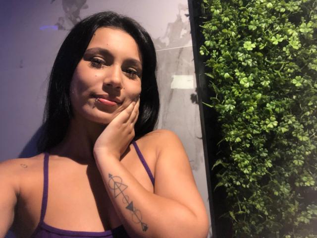 KataleyaMansson with long black hair, purple tank top, and tattoos on her right arm, touching her cheek with a slight smile, green plant on the right.