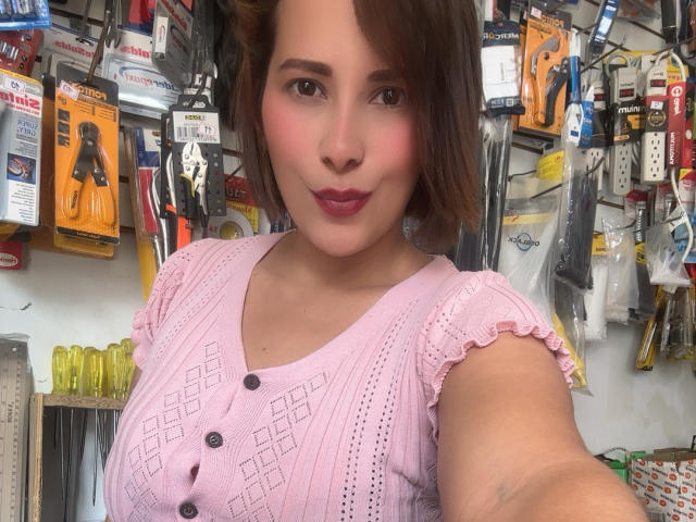 SaritaVelez draagt een roze top met knoopjes en korte mouwen, waardoor haar borsten zichtbaar zijn, en ze heeft volle, roze lippen. Ze staat in een winkel. Ze heeft kort, bruin haar, een lichte huid, donkere ogen en een subtiele blush. Zelfportret, lachend..