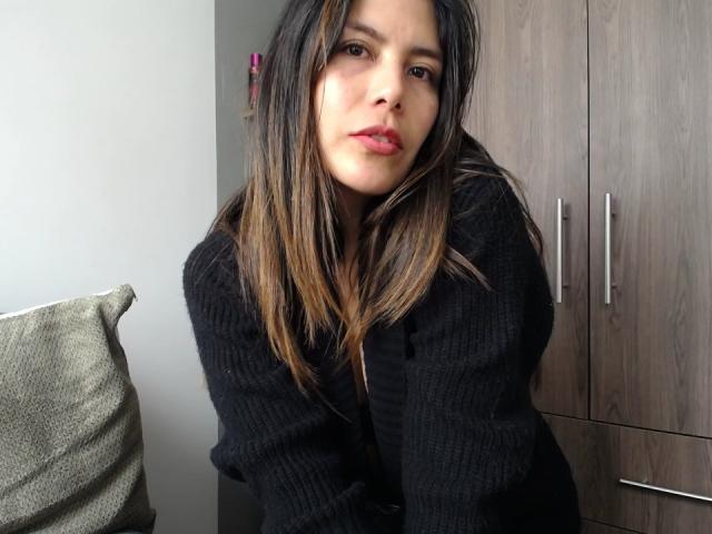 LeonaFey has medium brown skin, long dark brown hair with blonde highlights, and is wearing a black rib-knit sweater, leaning forward with a slightly pouty expression, showing her tits.