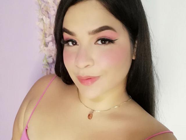 Antonela met roze oogschaduw, dikke wenkbrauwen, roze blush en roze lippenstift, draagt een roze top met spaghettibandjes, een ketting met een rode steen en heeft lang donker haar..