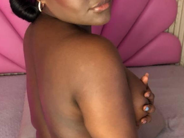 ScarlettHottiest, topless, dark brown skin, small round tits, covering one with hand, pink background, pink bed, pink heart-shaped pillow, black hair in bun, wearing hoop earrings.