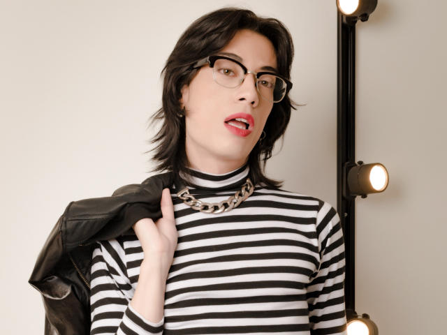 MilarHole with black-striped shirt, chain necklace, glasses, red lipstick, black jacket on shoulder, light bulbs in background, short dark hair, pale skin, looking slightly open-mouthed.