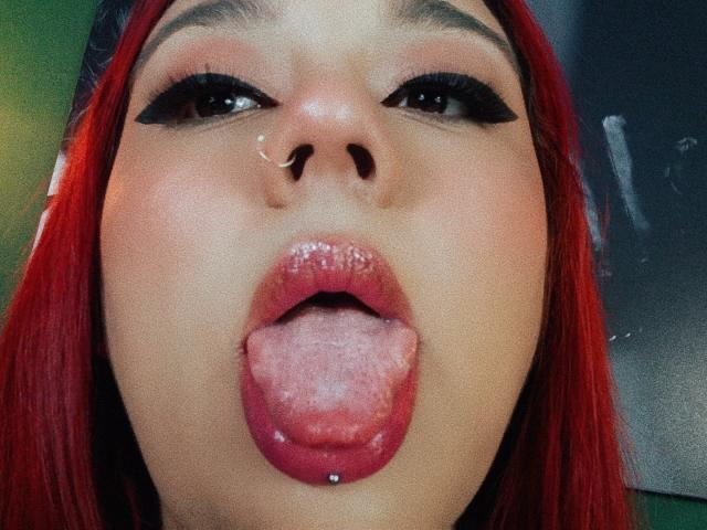 EmilyDom's face, tongue out, with a small nose piercing, red lipstick, black eyeliner, and red hair, showing a silver lip piercing. Her eyes are half-closed.