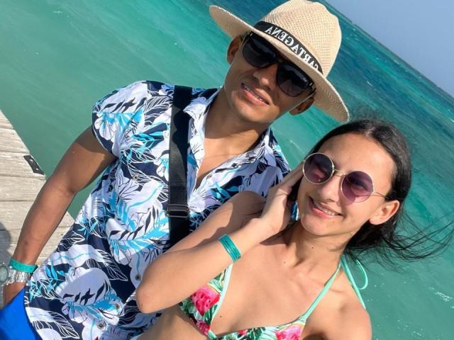 OsitaGab, con piel morena, vistiendo una camisa estampada, un sombrero de paja y gafas de sol, está de pie junto a OsitaGab, también con piel morena, con un bikini rosa estampado y gafas de sol redondas, frente a agua turquesa..