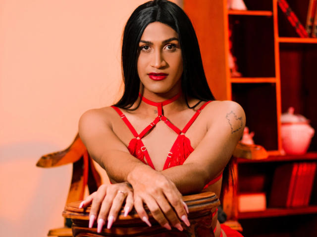 BarbaraPerri69 with long black hair, red lipstick, and red lace bra, arms on wooden chair, tattoo on left shoulder, red choker, red nails, looking at camera, smiling.
