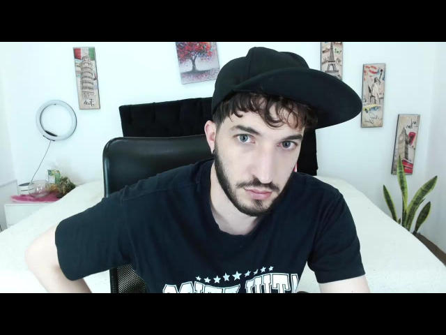 AllenMan, with a black cap and beard, leans forward, wearing a black t-shirt with white stars, showing a slightly confused or serious expression, in a bright room.