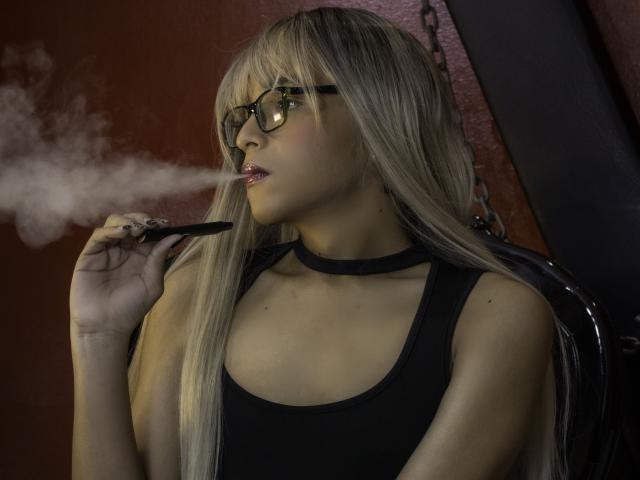 DannyHotz with long blonde hair, black glasses, and black tank top, smoking a joint, holding it near her pink lips, wearing a black choker, in a dark room.