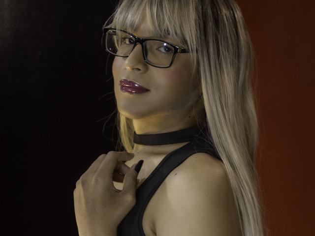 DannyHotz with long blonde hair, black glasses, shiny purple lipstick, black choker, and black sleeveless top, touching collarbone, smiling slightly.
