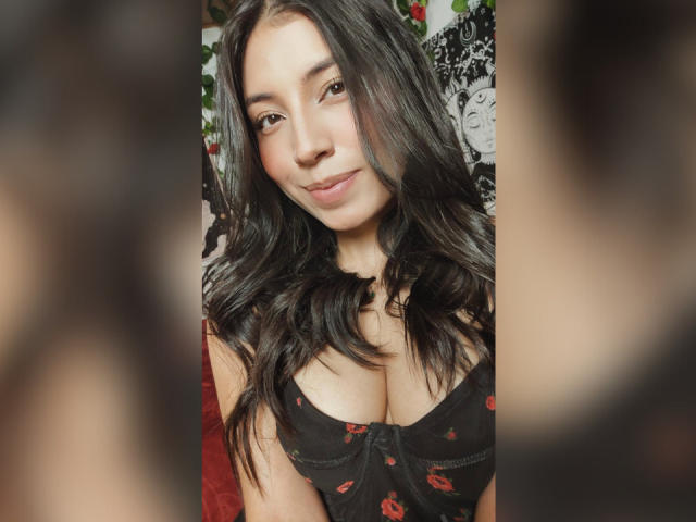 LorenDiaz with long black hair, smiling, wearing a black, red lip print, low-cut top, showing her big tits. Blurred faces in foreground. Soft lighting, cute.