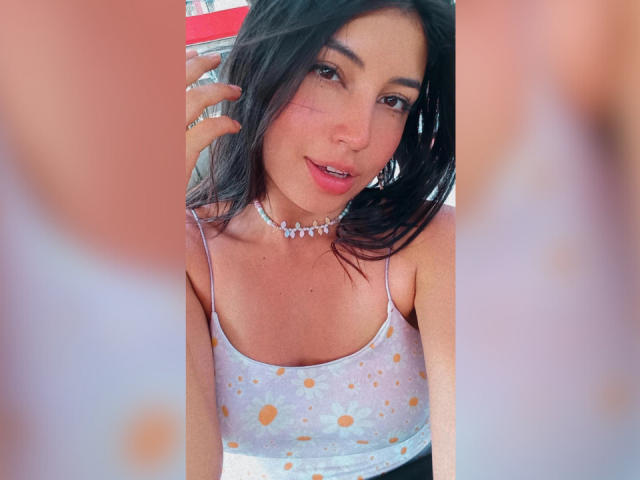 LorenDiaz with dark hair, wearing a white, gold-spotted tank top, and a jeweled choker, holding a red drink, with a slight smile, showing a small scar on her cheek.