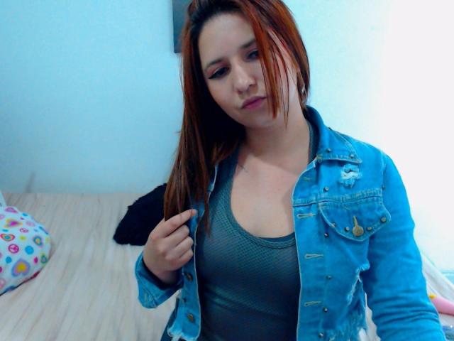 Kkatalina with red-brown hair, wearing a blue denim jacket over a gray tank top, holding her jacket collar, looking down with a slight frown. Boobs visible under top.