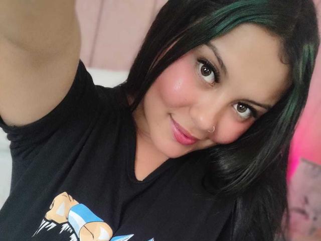 Camihotboobs with green highlights in her black hair, nose ring, pink blush, and black t-shirt, smiling at the camera with a slightly raised arm.