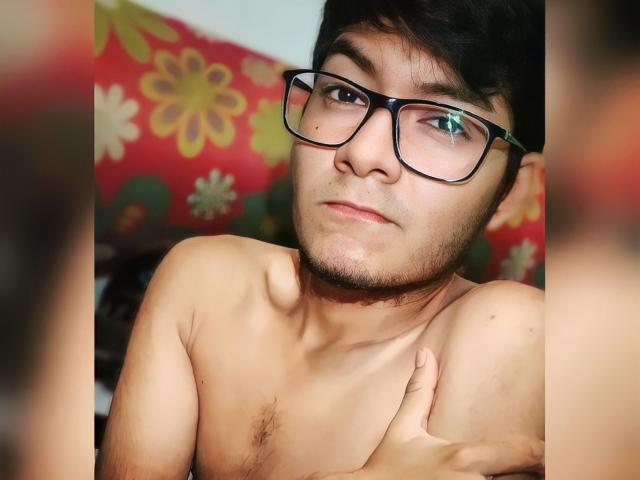 Lexden, sem camisa, usando óculos, com cabelo escuro e uma barba discreta, cobrindo o peito com uma mão, olhando para a câmera com uma expressão neutra..