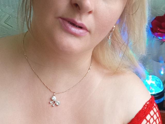 KelliRedhaer's face, showing pale skin, pink lips, blonde hair, gold necklace with a small diamond, and red fishnet top, exposing a bit of her tits.