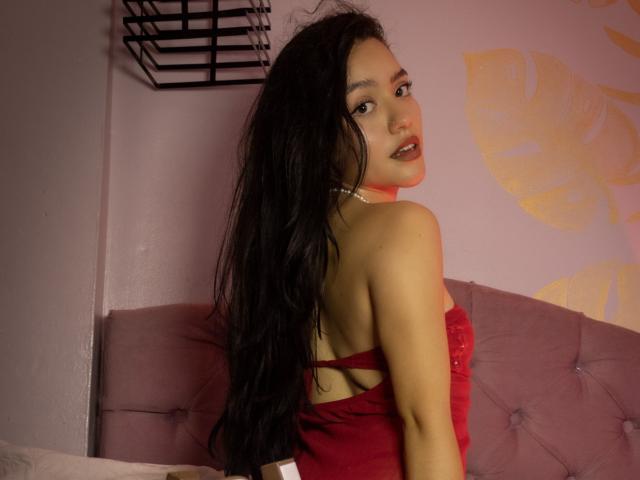 KateCrown, con i suoi lunghi capelli neri, guarda oltre la spalla, indossando un top rosso senza spalline che mette in mostra la sua schiena e rivela il suo piccolo e carino sedere. Ha una collana e un'aria sensuale..