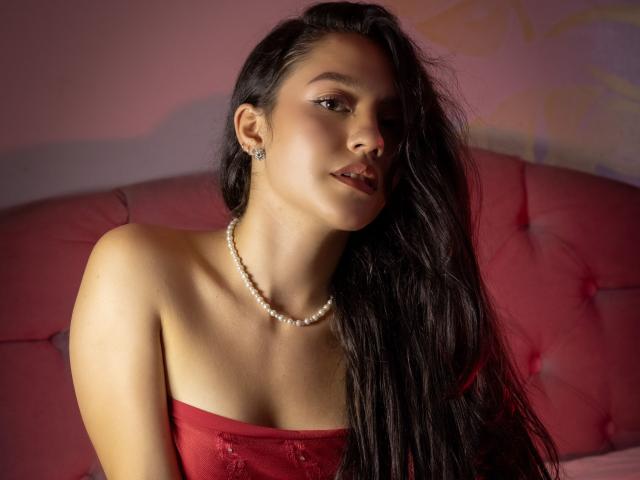 KateCrown with long black hair, wearing a red strapless top and pearl necklace, looking slightly to the side with a subtle smile, showing her tits and soft brown skin.