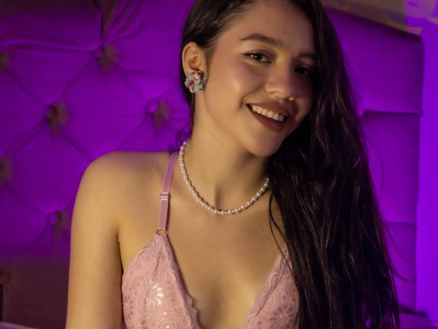 KateCrown, con lunghi capelli scuri, sorridente, indossa un reggiseno di pizzo rosa, una collana di perle e orecchini a forma di farfalla, ha un seno carino e un sedere carino, illuminata da una luce viola..