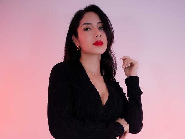 ArianaRogers with dark wavy hair, red lipstick, black V-neck sweater, slight cleavage, right hand near face, pink-purple gradient background, looking confident.