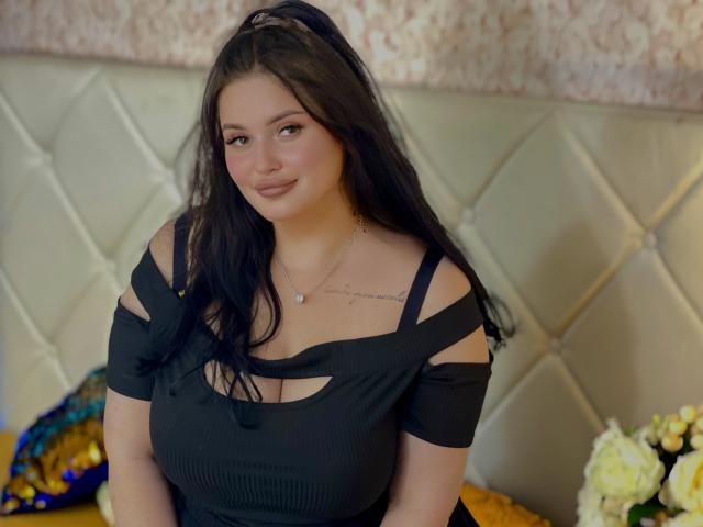 MonaSteele, con cabello negro largo, blusa negra con un hombro descubierto que muestra sus senos, una sonrisa sutil, un tatuaje en el pecho derecho, un collar, sentada en una cama con flores blancas y amarillas..