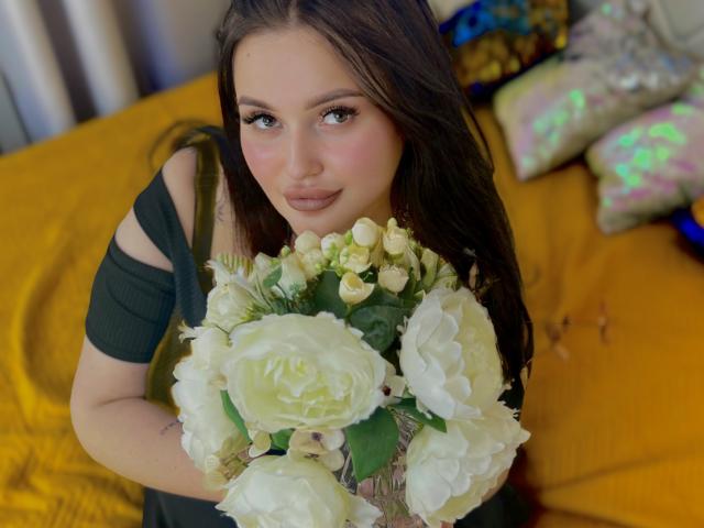 MonaSteele avec des roses blanches, tenant un bouquet, yeux verts, cheveux bruns, haut noir sans manches, sourire doux, lit jaune, oreillers brillants en arrière-plan..