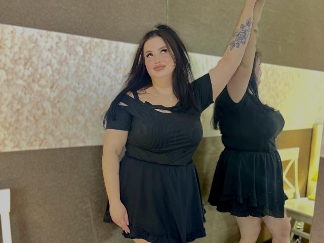MonaSteele con vestido negro, brazo levantado, tatuaje en el brazo izquierdo, busto grande, cabello castaño, sonriendo, de pie junto a un espejo, que refleja su imagen..