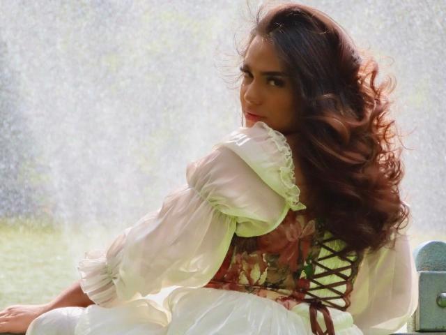 LiliithBuckland with curly brown hair, white puffed-sleeve dress, and floral corset, sitting on a green bench, looking back with a sultry expression, soft light, outdoor setting.